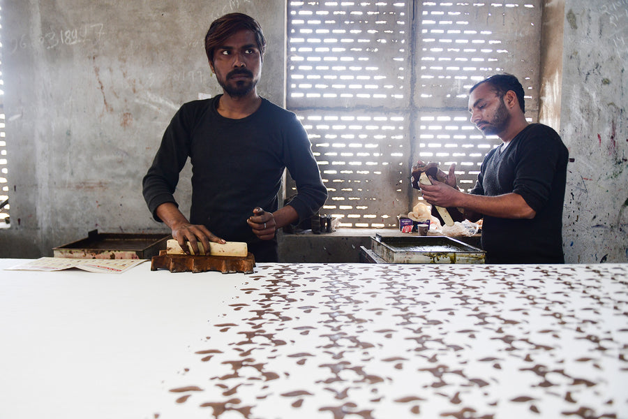Two men print fabric by hand using wooden blocks as sun streams in.