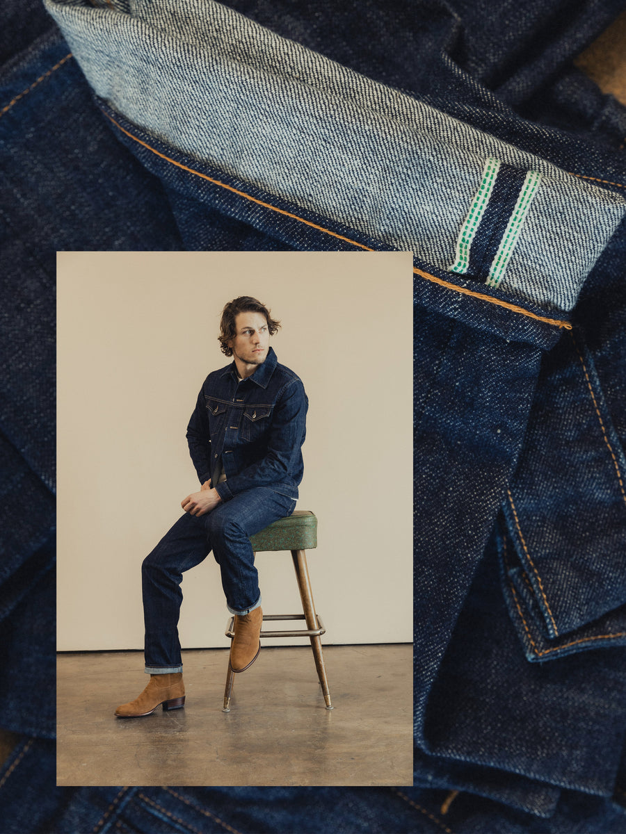 A man in a denim jacket and jeans sits on a stool.