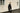 A man in a black shirt stands against a wall.