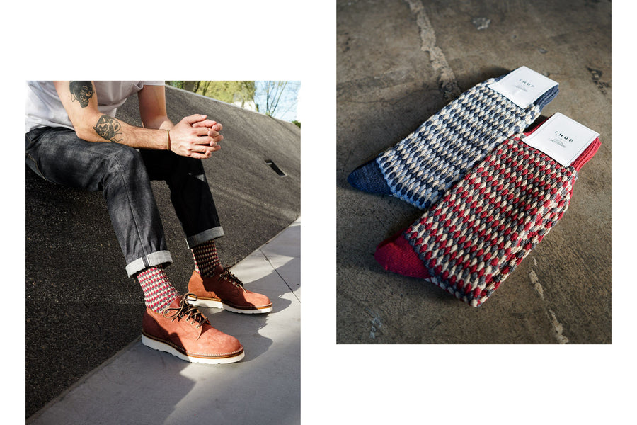 A man sits on a sloped surface showing off the red patterned Chup socks paired with blue jeans and Viberg oxfords.
