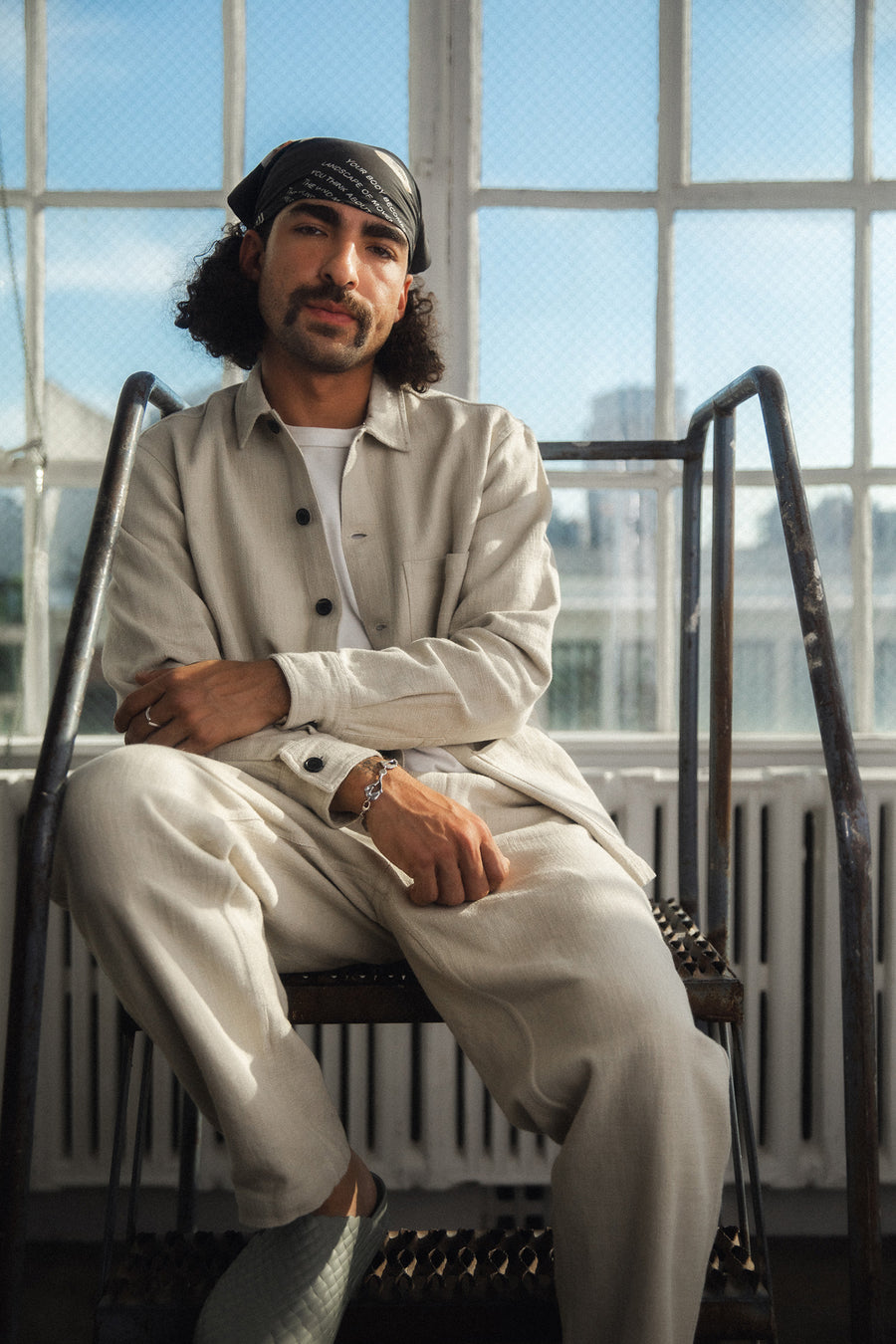 A man in a white linen jacket and pants with grey clogs sits on a metal staircase.