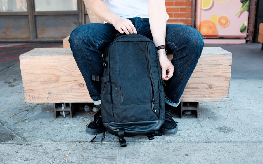 A front view of the black backpack while a model holds it between his legs while sitting on a bench.