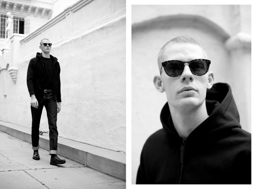 A man stands against a white wall in a 3sixteen hoodie and jeans.