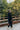 A man in a black wool coat walks across a bridge in a park.