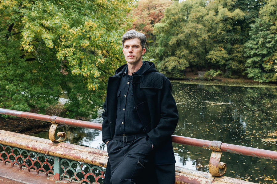 A man in a black duffel coat stands on a bridge in a park.