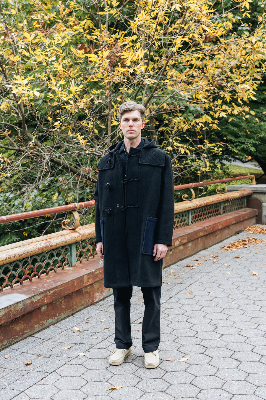 A man in a black wool coat stands on a bridge.