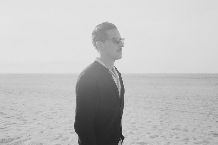 A man in a black cardigan and a button down looks across a beach.