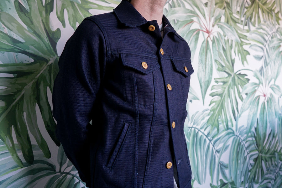 The model stands in front of a greenery-filled wall wearing the Type 3s Jacket buttoned all the way up.