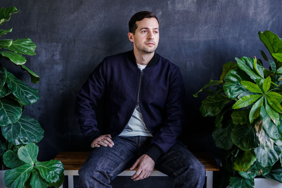 A man sits in a room surrounded by plants wearing the 3sixteen Stadium jacket in Panama Cloth.