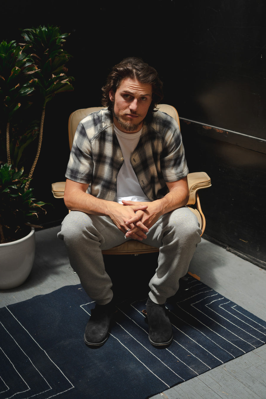A young man in a plaid baseball shirt and grey sweatpants sits on the edge of a chair.