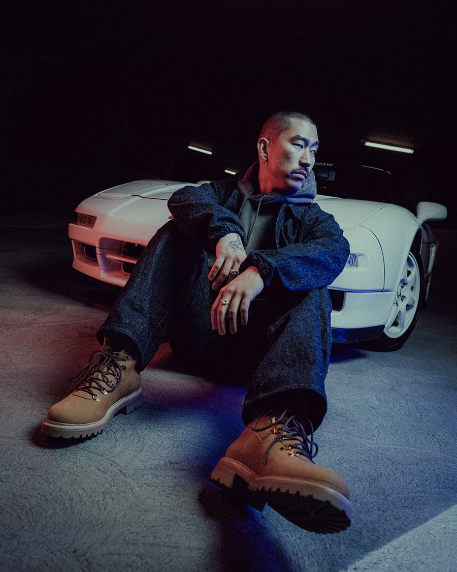 A man in a denim coach's jacket and jeans sits in front of a white sports car.