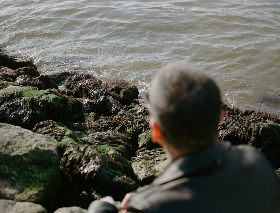 An out of focus man sits off frame while gentle waves wash up onto rocks on the shore.