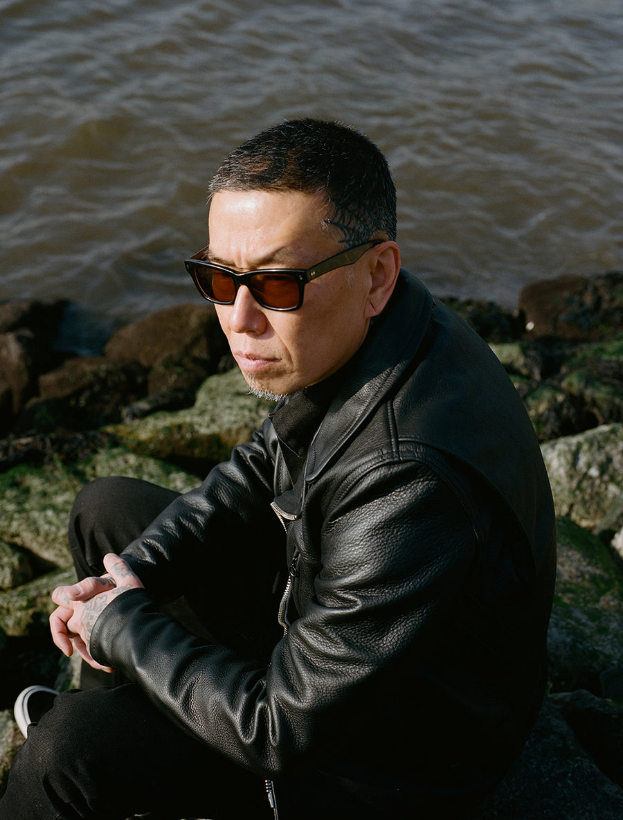 A man in a black leather jacket sits upon mossy rocks on the shore.