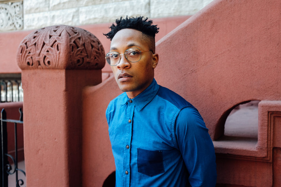 A man wears an indigo long sleeve button down shirt in front of a red stoop.