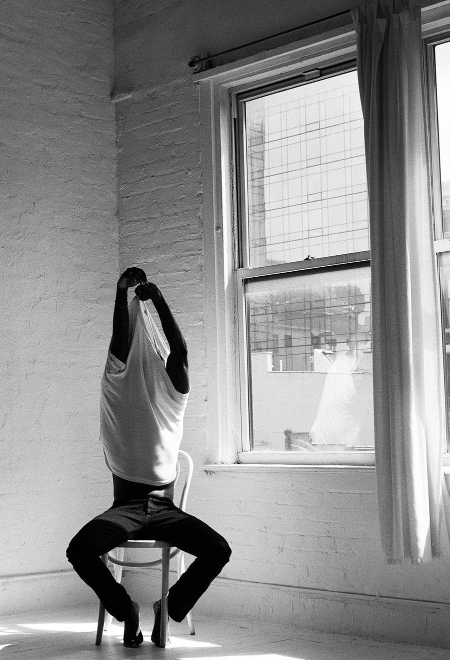 The model pulls a 3sixteen white tee off his body while seated in a chair in a white room.