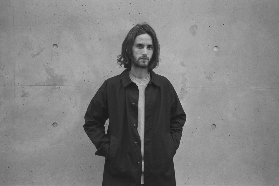 A man in a black mac coat stands against a wall.