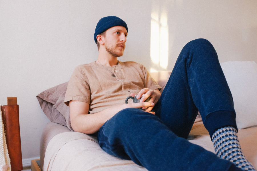 The model holds a coffee mug in bed while wearing the indigo sweatpants.