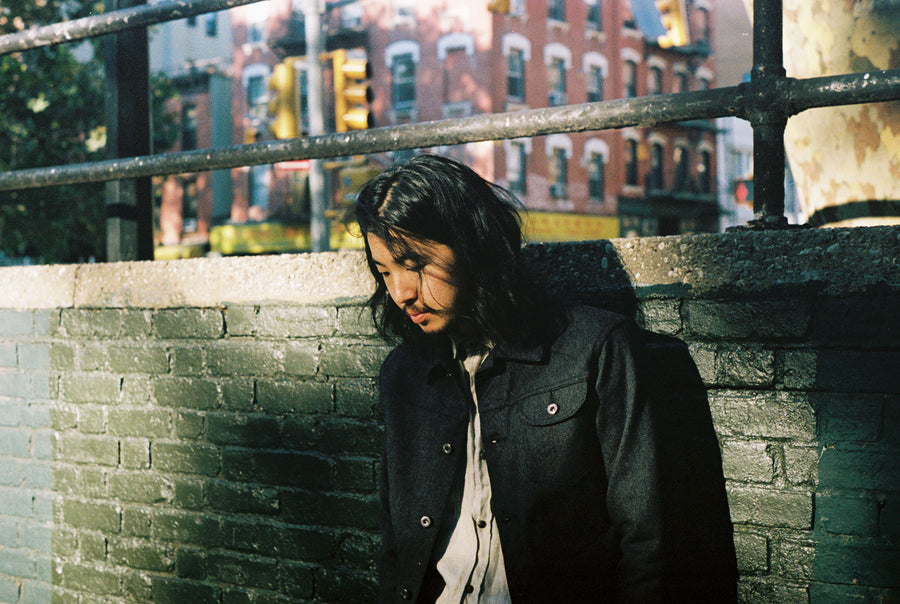 The model, wearing a 3sixteen Ranch Jacket, leans against a green brick wall and looks down.