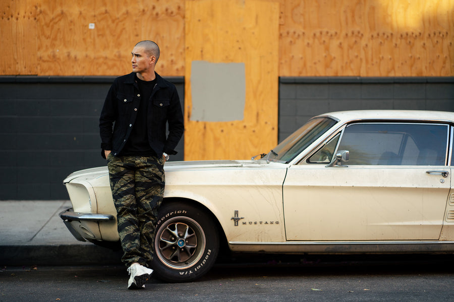 Man leans against a vintage Mustang in a black jacket and camouflage pants