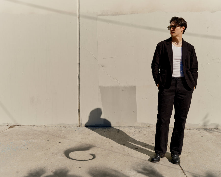 A man in a black suit and white tank stands by a wall.