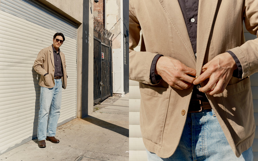 A man in a khaki blazer and light wash jeans stands in front of a gate.