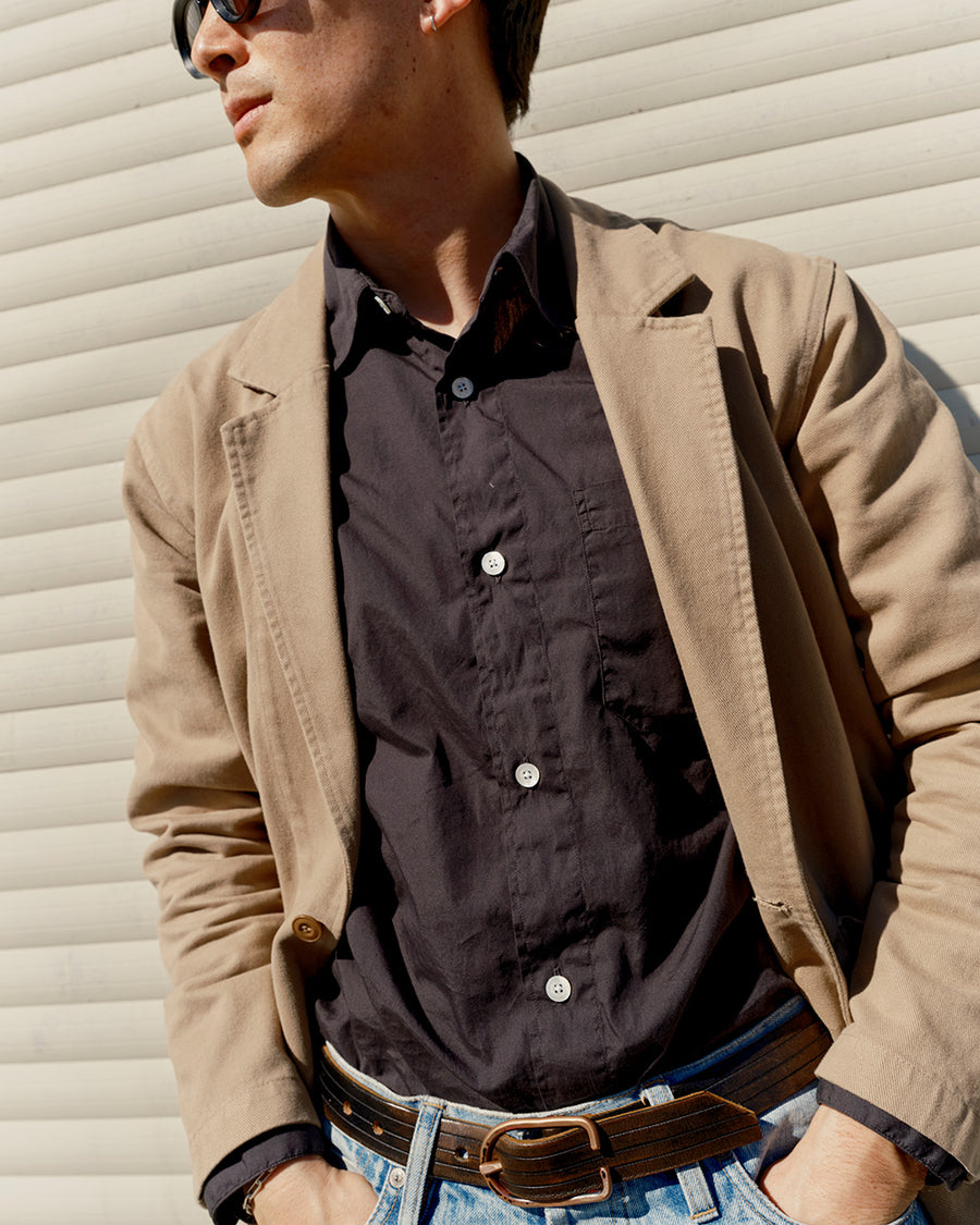 A man in a khaki blazer and black oxford stands outside.