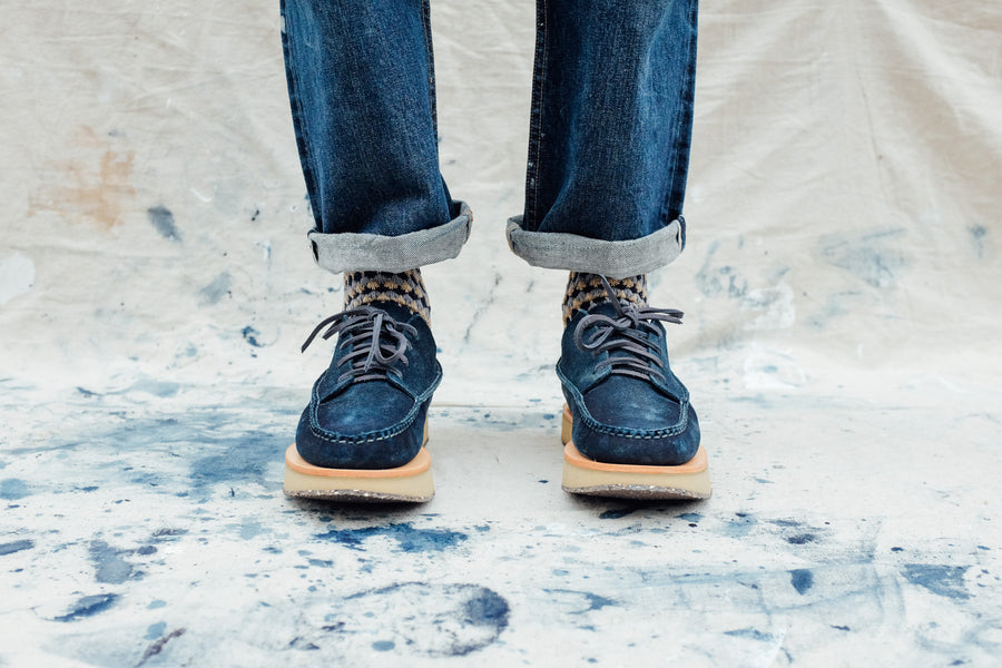 Two feet clad in blue moccasins stand on an indigo-stained cloth.